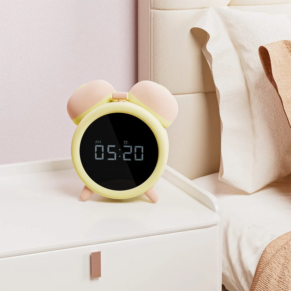 Product detail image for the Naughty Clock Vibrator Sonaire, featuring a round digital alarm clock with a pink and yellow twin-bell design placed on a white bedside table. The display reads “AM 05:20” and includes a small icon, suggesting multifunctional features. The bedroom setting includes soft pink walls and beige bedding, creating a warm, intimate atmosphere. The composition emphasizes Sonaire’s blend of classic aesthetics and modern functionality, reinforcing its discreet dual-purpose as a luxury vibrator and a stylish bedside clock.